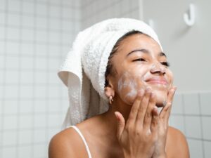 Woman in tiled bathroom exfoliating her face with a towel around her head