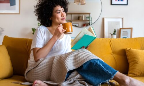 Woman on a couch with a book and mug and blanket