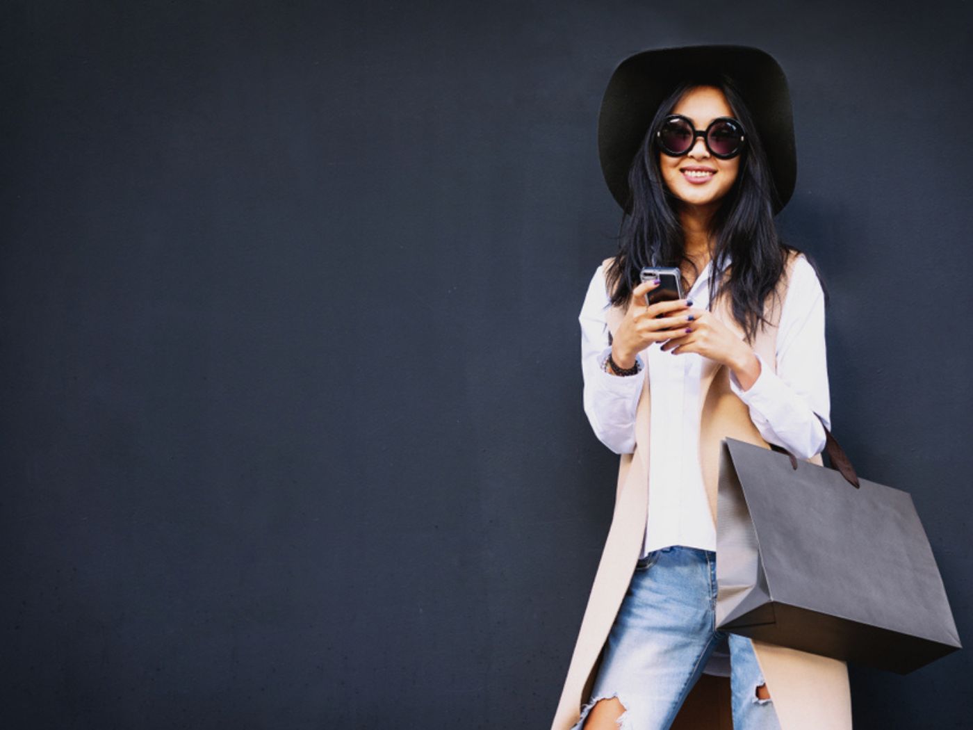 Woman with dark hair and a large hat with sunglasses carries her phone and a shopping bag