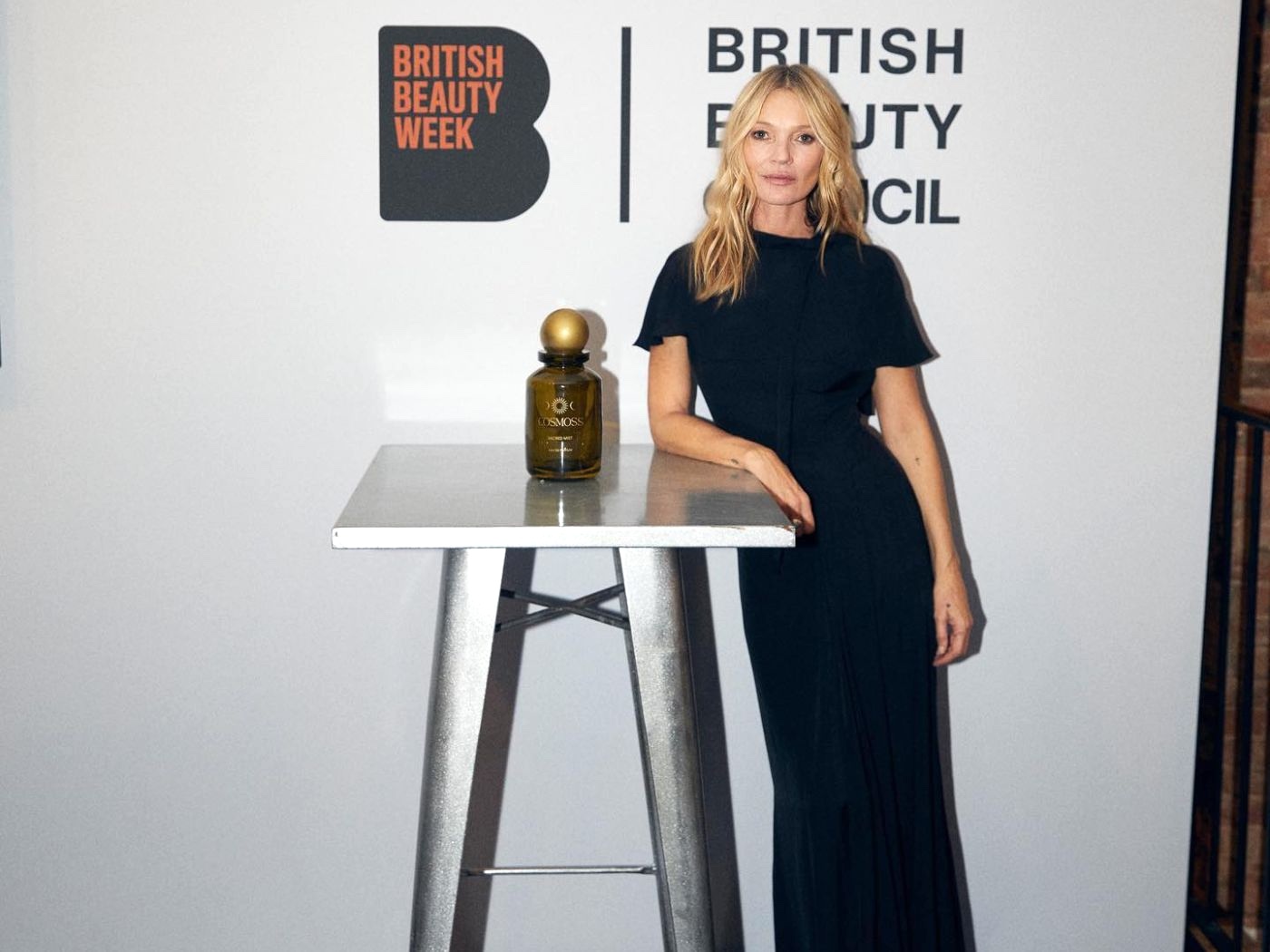 Kate Moss, with long blonde hair worn down and wearing a long black dress poses by a tall table with a bottle of beauty product. British Beauty Council logo on wall behind her.