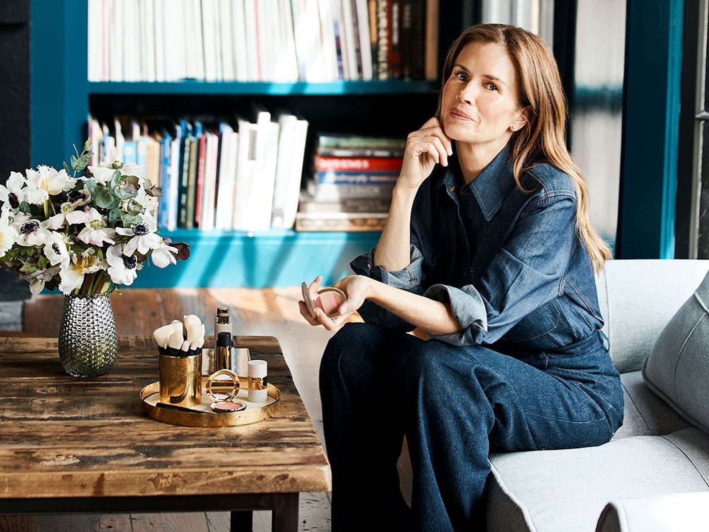 Woman wearing a denim shirt and jeans is seated on a couch by a table of make-up, holding a make-up compact in one hand.