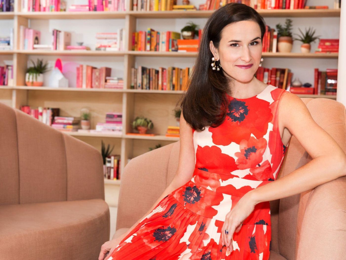 Woman in a red floral sleeveless dress sitting in a chair in front of a bookshelf.
