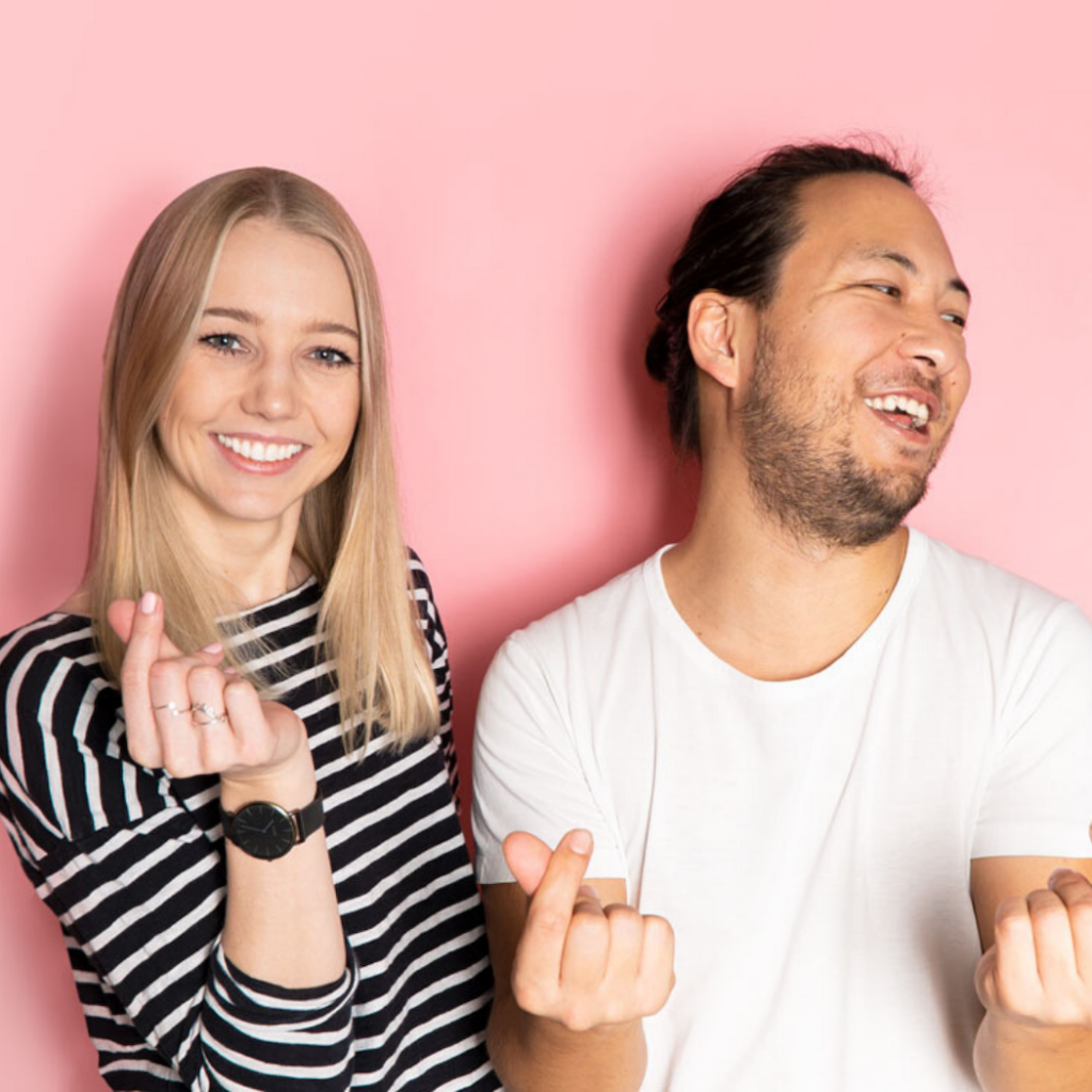 A woman with long blonde hair in a striped black and white top stands next to a man with dark hair in a white t-shirt, against a pink background. They are clicking their fingers and laughing.