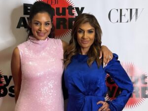 Two women stand side by side, one in a pink sequinned sleeveless dress and the other in a long-sleeved blue silk dress.