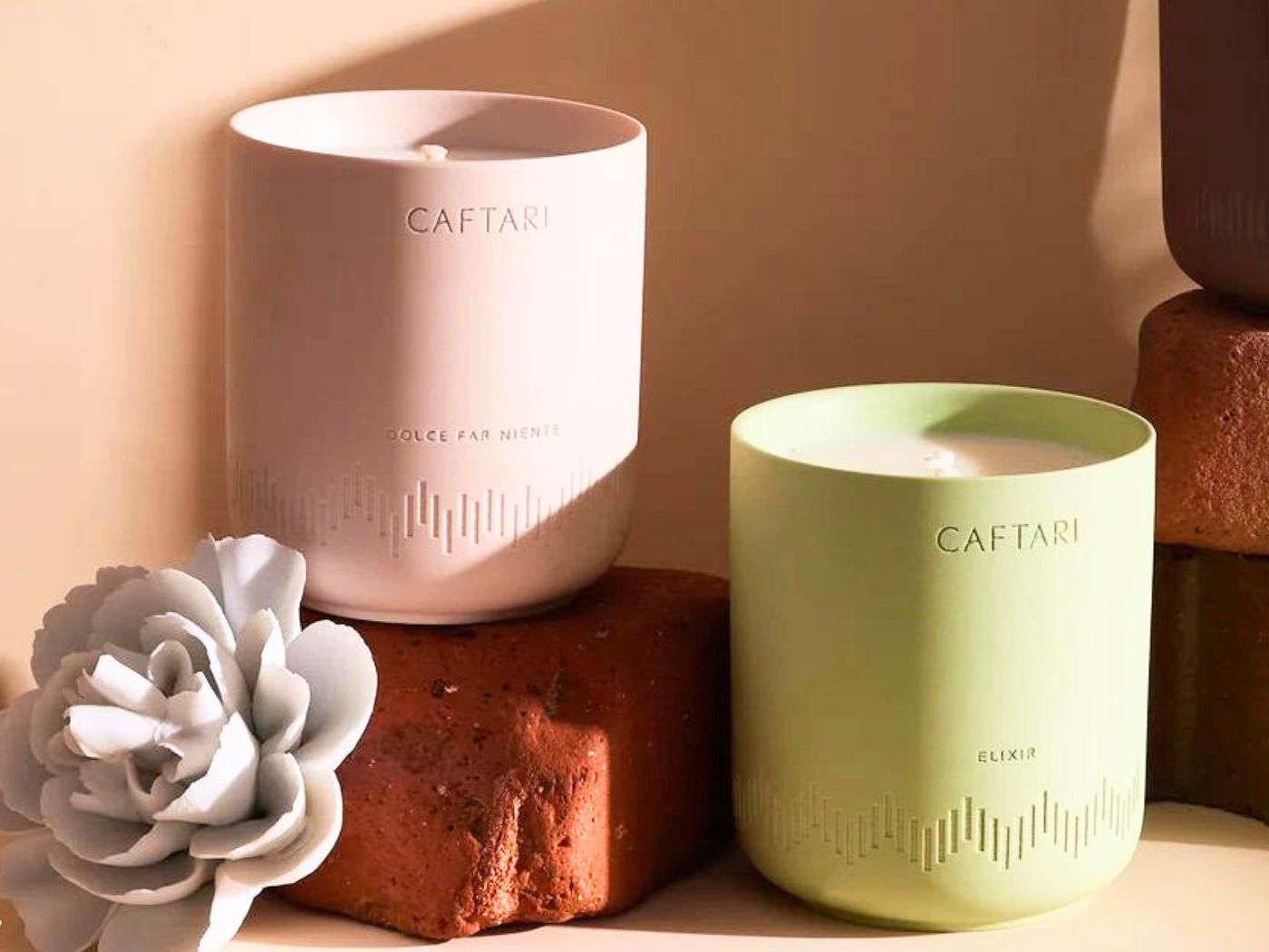 Two votive candles, pale pink and soft green, are displayed with red rocks and a white ceramic flower.