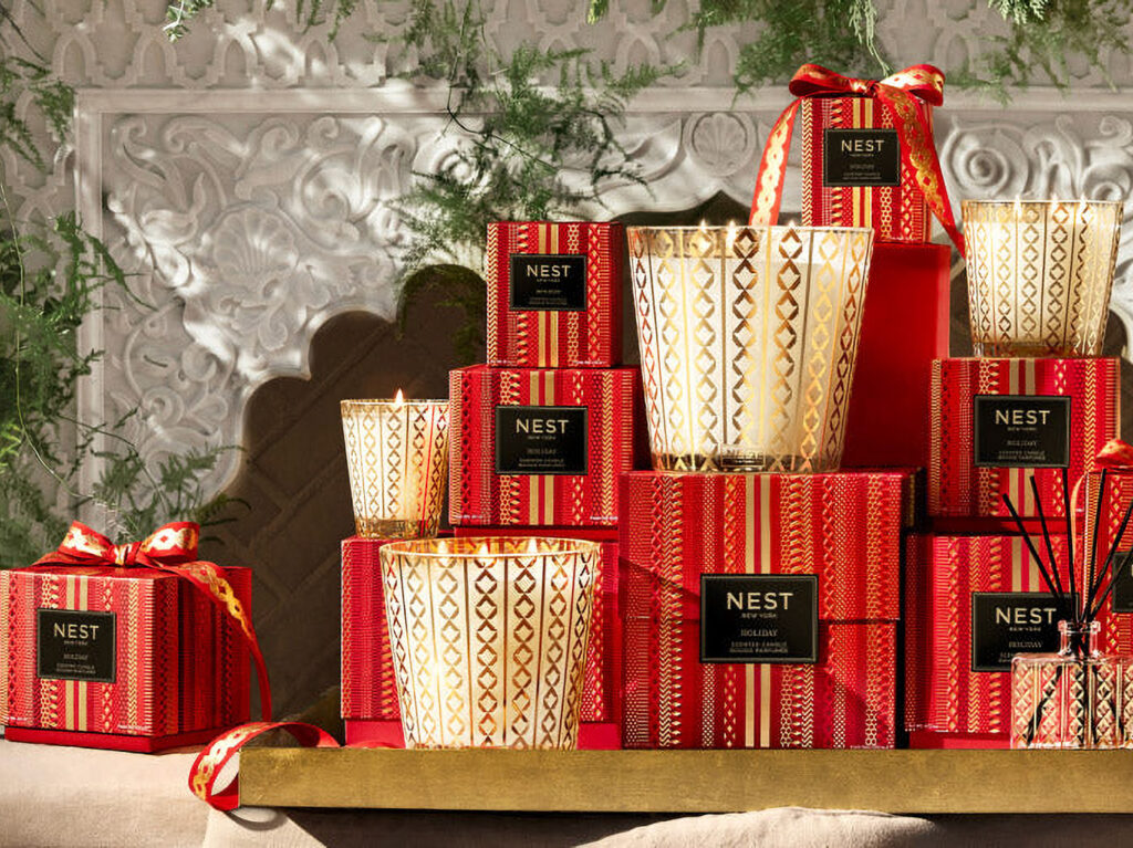 An array of scented candles in glass jars with gold details, and their red, black and gold boxes, are displayed on a gold shelf with festive greenery in the background.