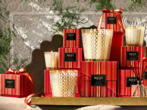 An array of scented candles in glass jars with gold details, and their red, black and gold boxes, are displayed on a gold shelf with festive greenery in the background.