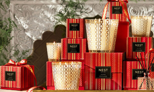 An array of scented candles in glass jars with gold details, and their red, black and gold boxes, are displayed on a gold shelf with festive greenery in the background.
