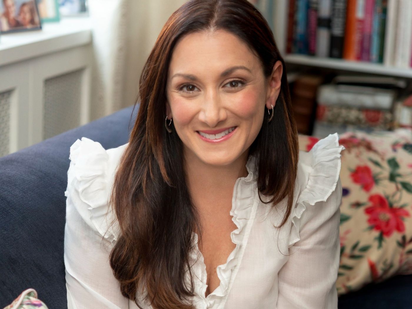 A woman in a white blouse sits smiling on a next to a floral cushion and a bookshelf.