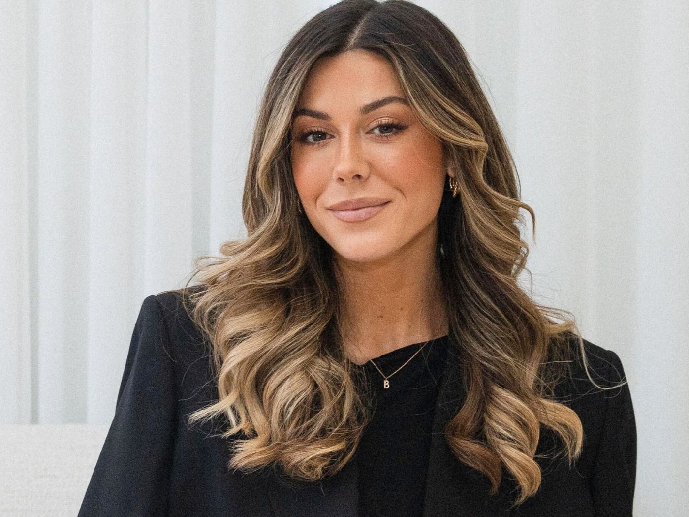 A woman with long, centre-parted hair with waves, and glowing make-up, wearing a black blazer and sweater.