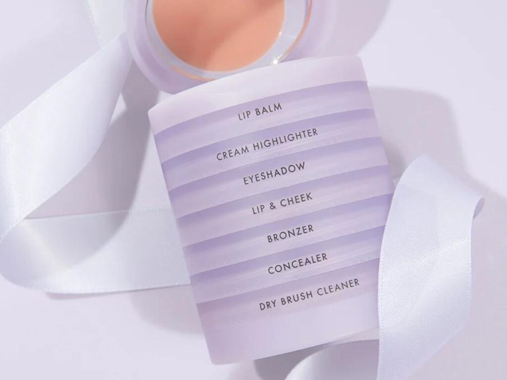 A stack of white make-up jars, clicked together, lies on top of a white unfurled gift ribbon.