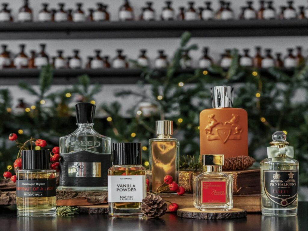 A selection of perfume bottles arranged on a shelf, with festive foliage and fairy lights and apothecary style bottles on shelves in the background.
