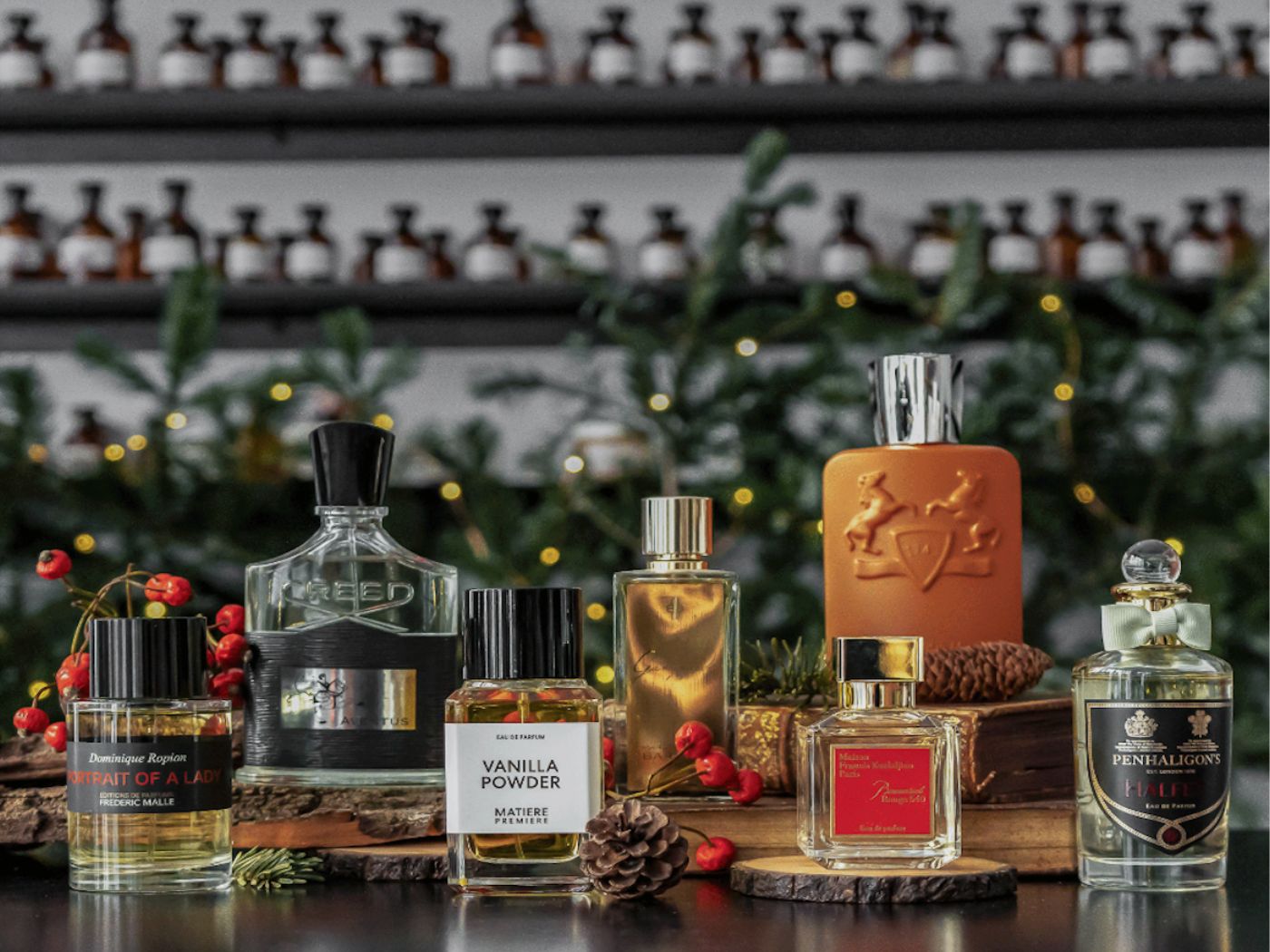 A selection of perfume bottles arranged on a shelf, with festive foliage and fairy lights and apothecary style bottles on shelves in the background.