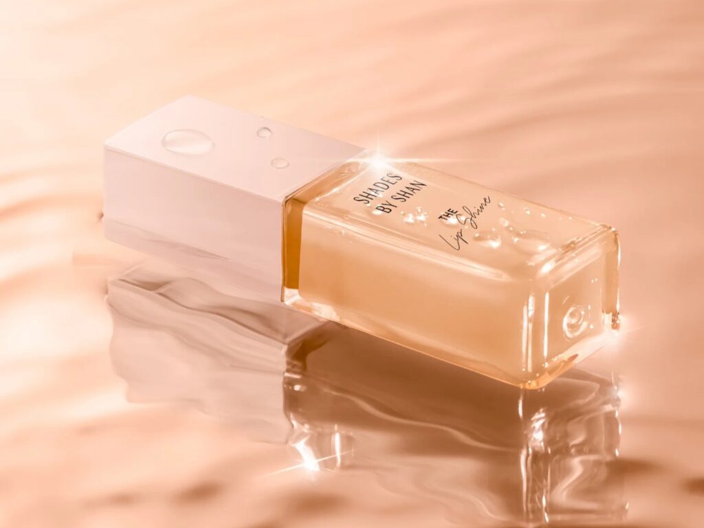 A tube of peach-coloured lip gloss is laid on a matching, rippling surface.