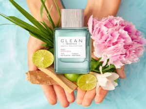 A blue glass bottle of perfume is held in model's hands, with a peony bloom and a selection of fruits and plants.