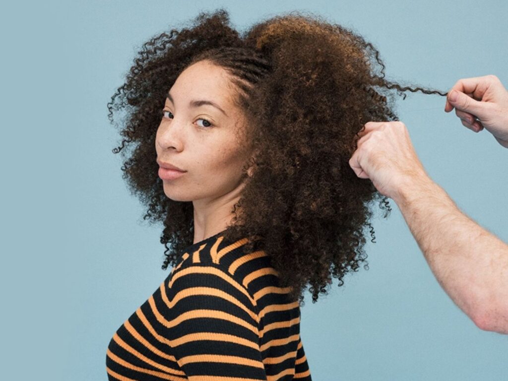 Model with textured hair wearing and orange and black striped t-shirt has her hair styled against a blue background.