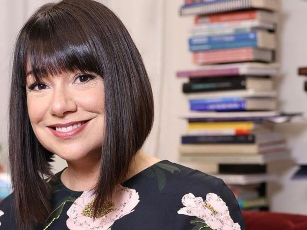 Woman with jaw-length, dark brown hair smiling, with a stack of books in the background.