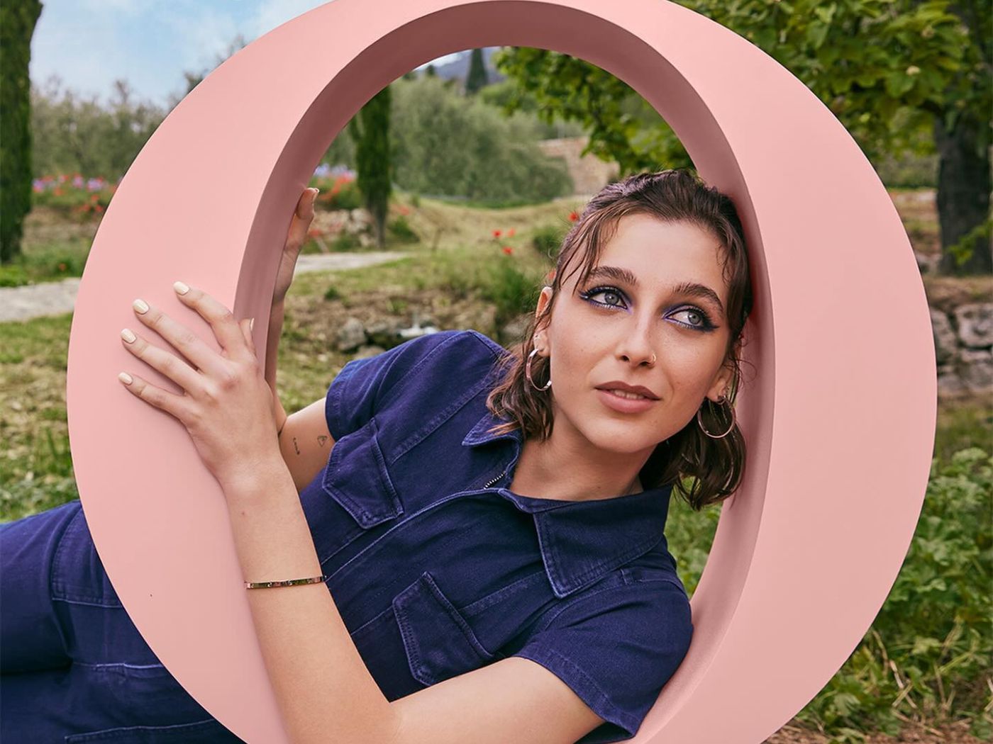 Girl in denim jumpsuit poses inside a giant pink letter "O" on grass.