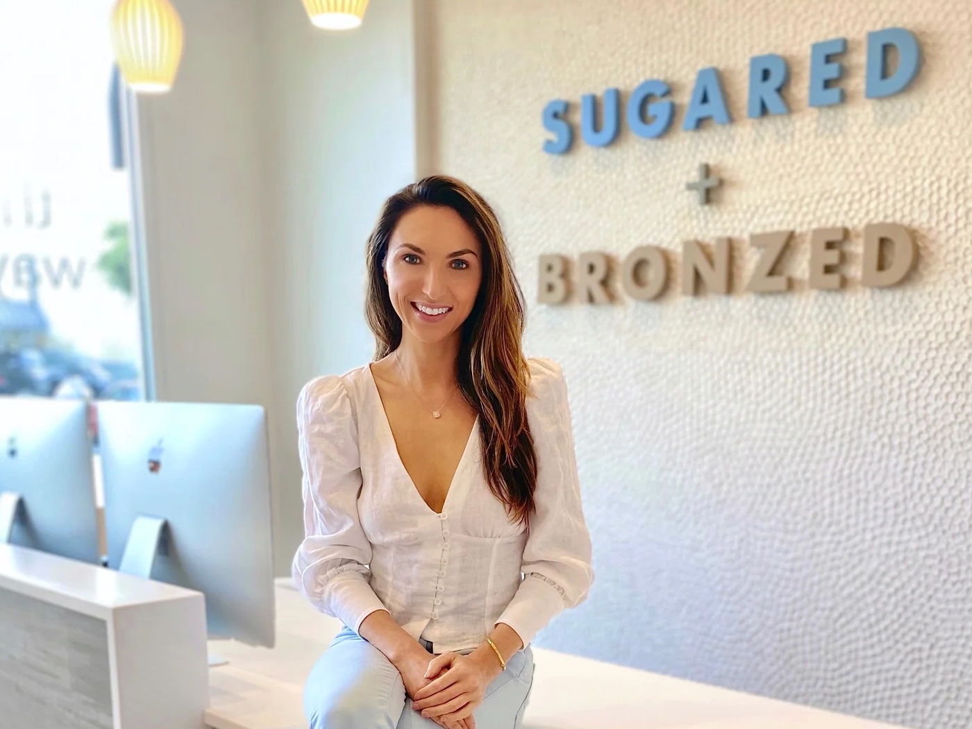 Woman wearing jeans and a white blouse sits on front desk of salon reception area, with Sugared + Bronzed logo on wall behind her.