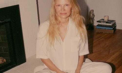 Pamela Anderson sits cross-legged in white pyjamas on a wooden floor in front of a fireplace.