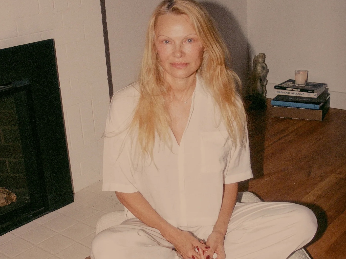 Pamela Anderson sits cross-legged in white pyjamas on a wooden floor in front of a fireplace.
