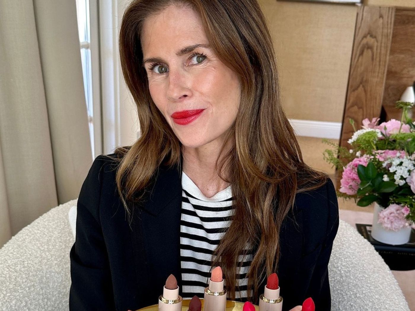 Woman in black blazer and striped black and white t shirt wearing red lipstick, seated holding a tray of oversized lipsticks in different shades.