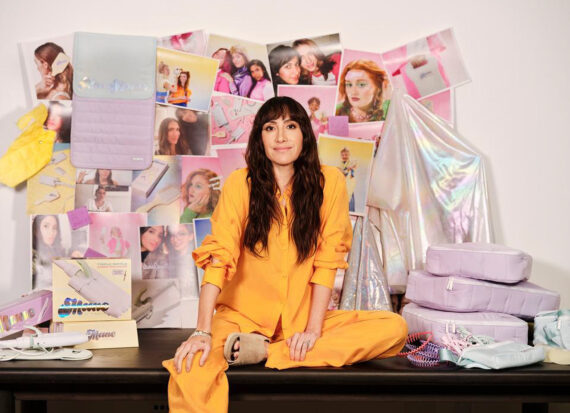 Woman sitting on a desk wearing a bright yellow blouse and pants. pinboard with lots of photos behind her.