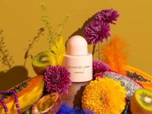 A pale pink opaque bottle of perfume, positioned among brightly coloured fruits and flowers.