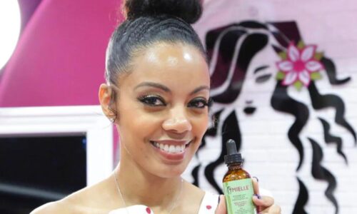 Monique Rodriguez stands smiling, holding a Mielle Organics product in a brown apothecary bottle, with a green label.