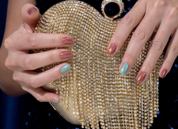 Hands holding a beaded sparkling handbag, with nails painted alternately with teal and brown metallic polish.