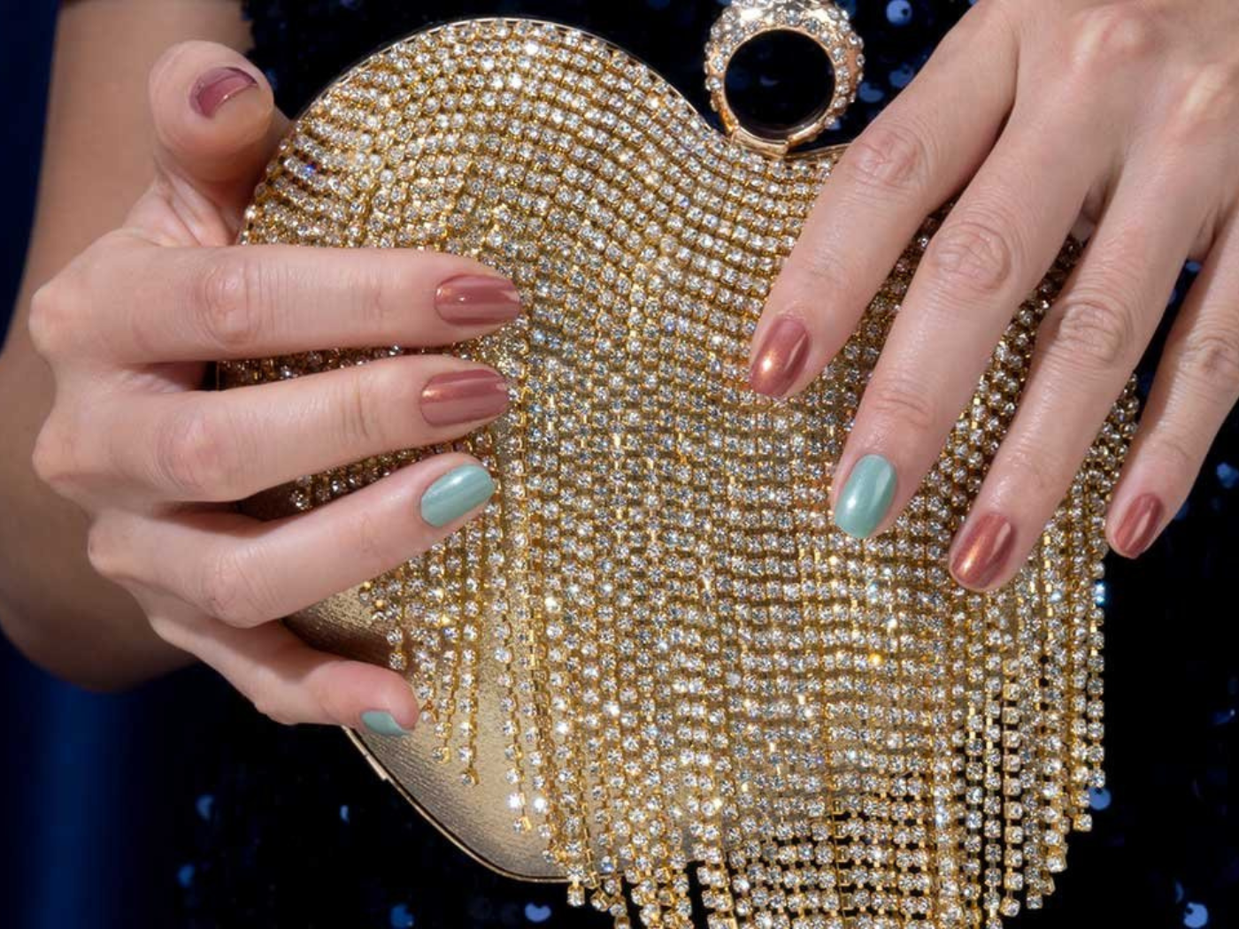 Hands holding a beaded sparkling handbag, with nails painted alternately with teal and brown metallic polish.