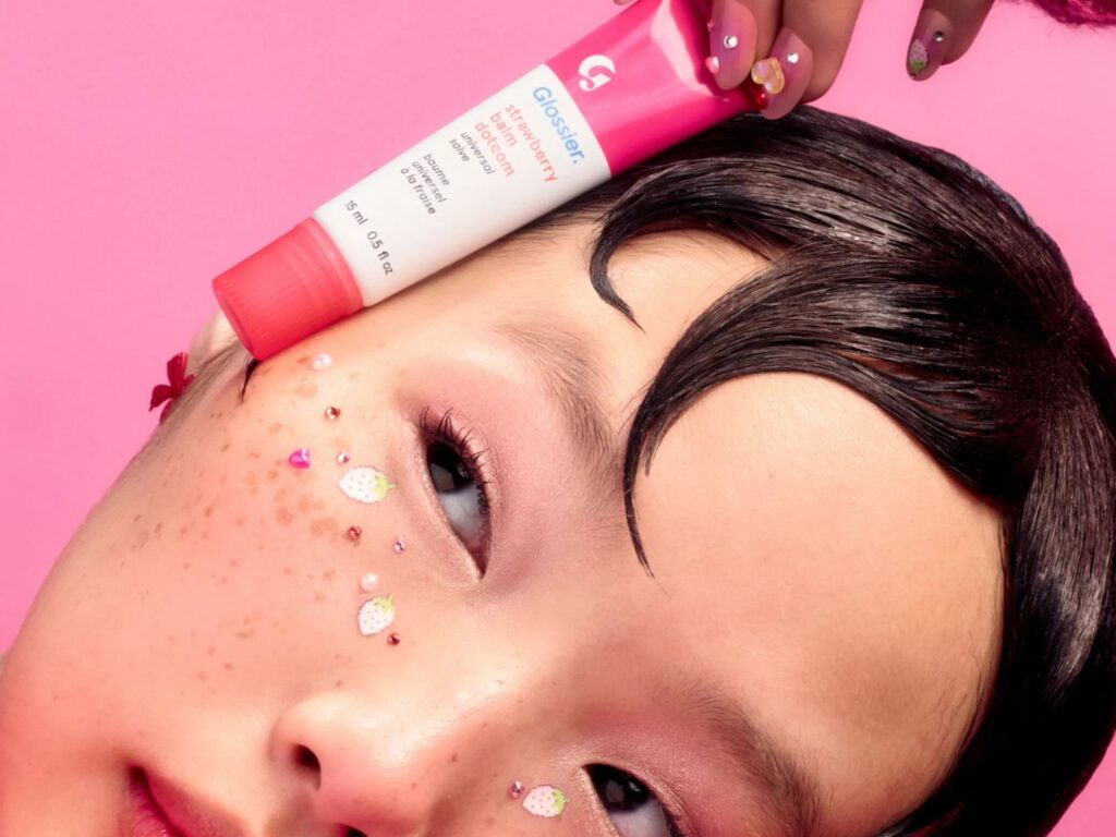 Girl with stick-on sparkly freckles holds a tube of pink Glossier lip balm gloss against her face.