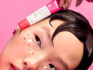 Girl with stick-on sparkly freckles holds a tube of pink Glossier lip balm gloss against her face.
