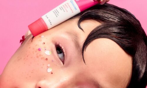Girl with stick-on sparkly freckles holds a tube of pink Glossier lip balm gloss against her face.