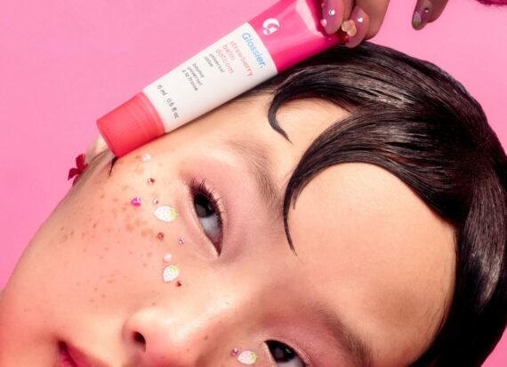 Girl with stick-on sparkly freckles holds a tube of pink Glossier lip balm gloss against her face.