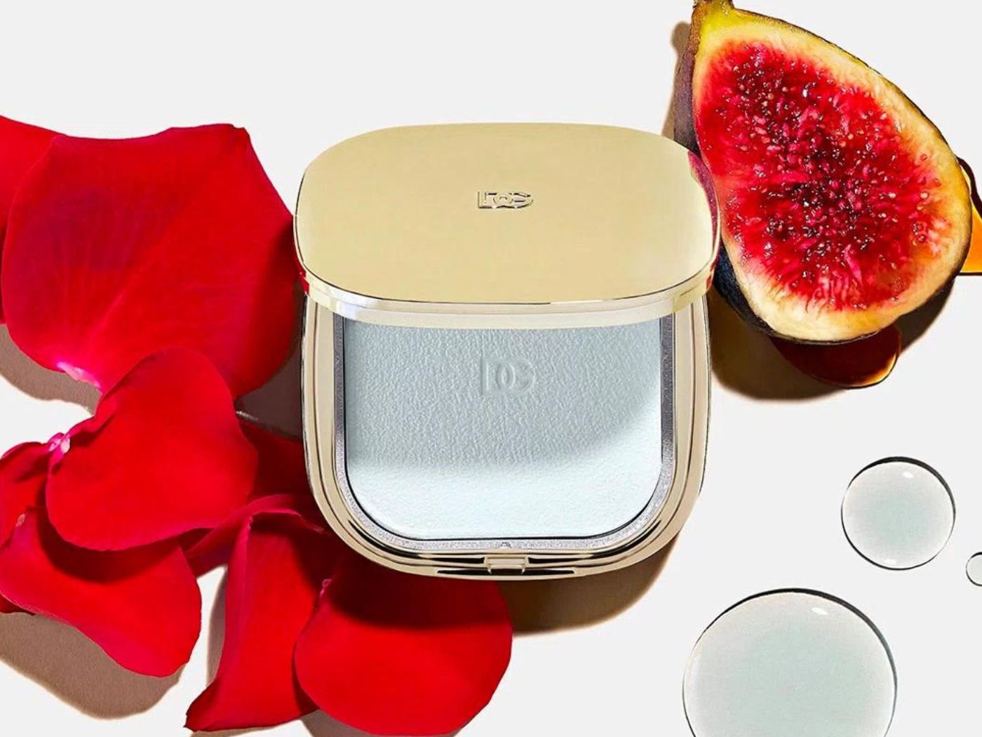 A pale blue eyeshadow compact with gold lid, surrounded by a fig and rose petals