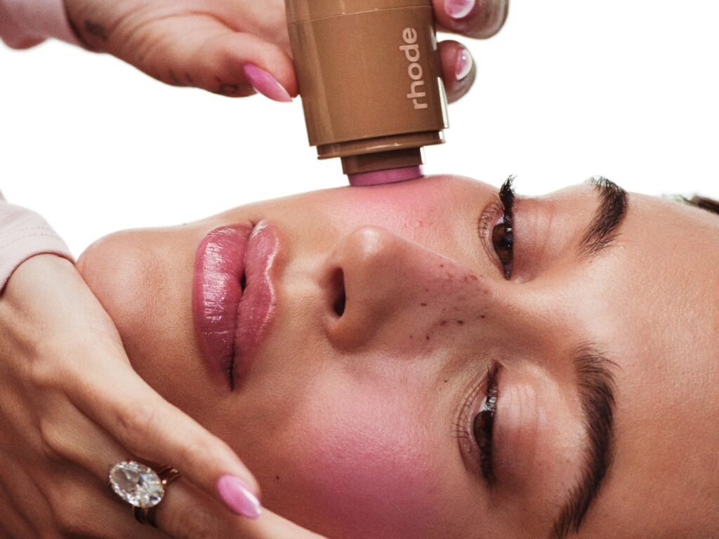 Hailey Bieber applies a blush stick to her cheeks.