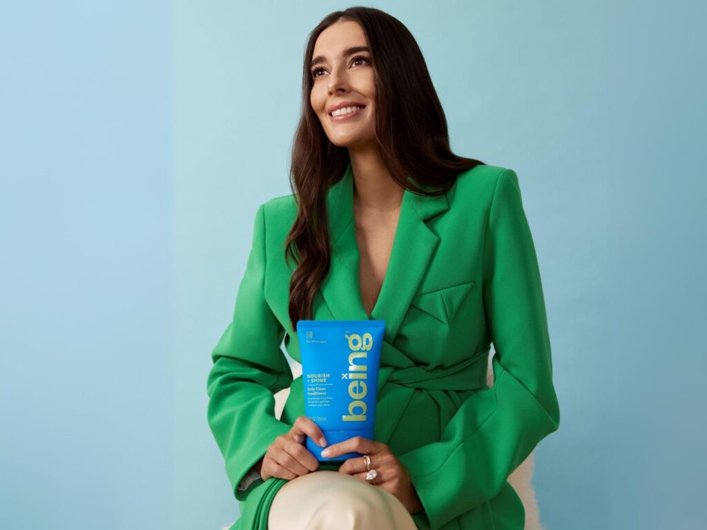 Woman in a green blazer holding a bright blue tube of product labelled "being"