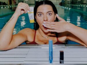 Olympic swimmer Anastasia "Tas" Pagonis in a swimming pool, holding her hand to her mouth and, in one hand, a tube of elf cream and, in front of her poolside, two elf makeup products.