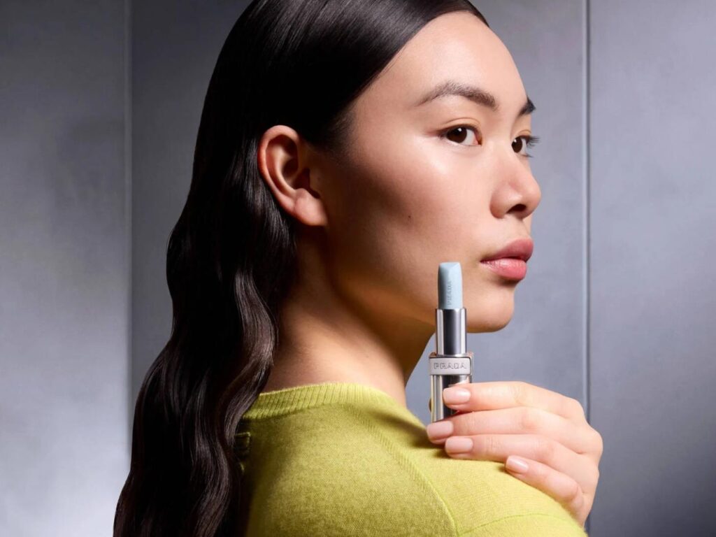 Model in lime green sweater holds a pale blue lipstick with PRADA logo