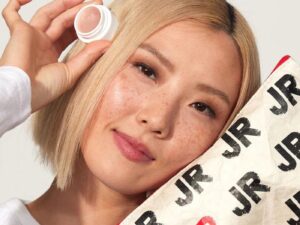 Woman holding a mini white pot containing a shimmering balm, and holding a makeup bag with the initials JR on it