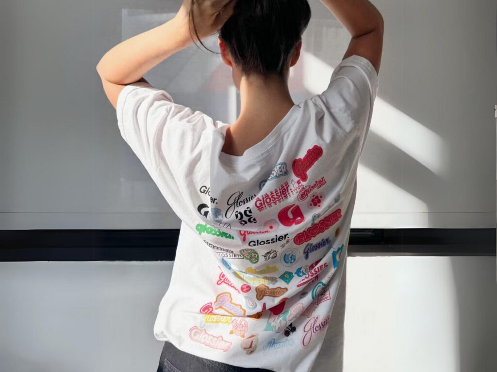 Photo shows a model wearing a white-t shirt, in rear view, covered with Glossier sticker images