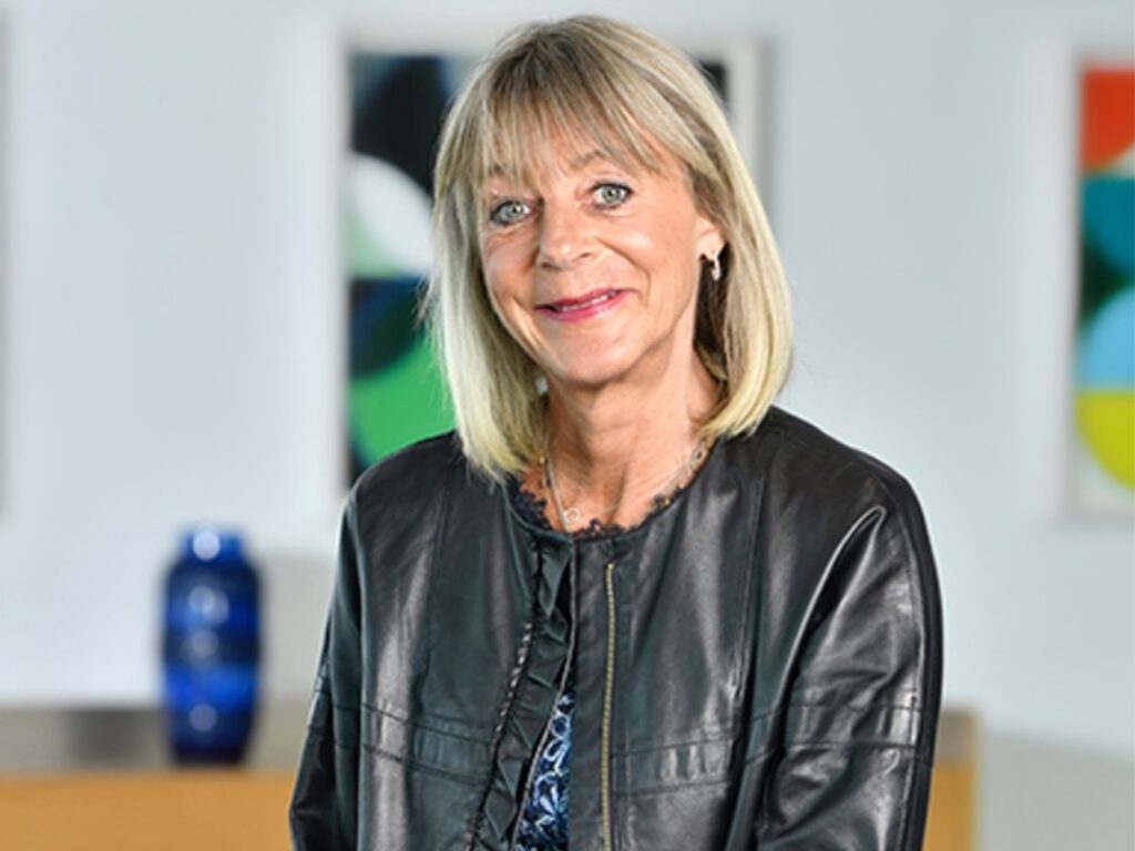 Photo of Sue Fox wearing a leather jacket in an office with brightly coloured artwork on the wall behind her