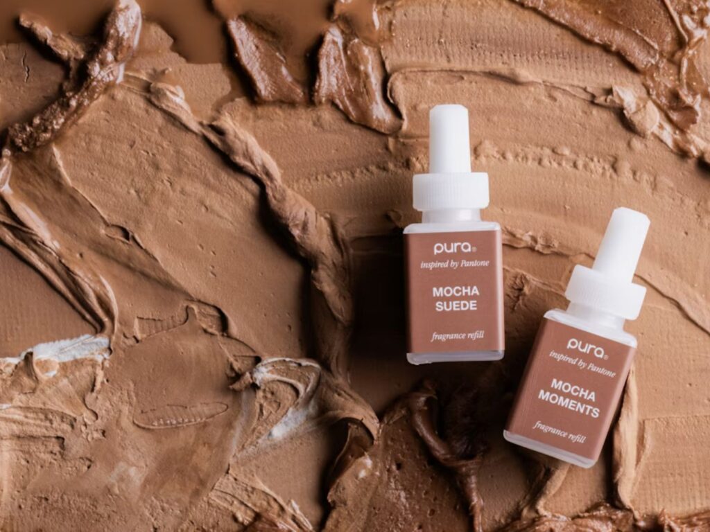 Two mocha-coloured Pura fragrance bottles displayed against a palette of mocha-coloured, creamy makeup swirls