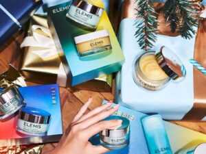 A selection of colourful Elemis products and packages under a Christmas tree, with a hand holding one jar.