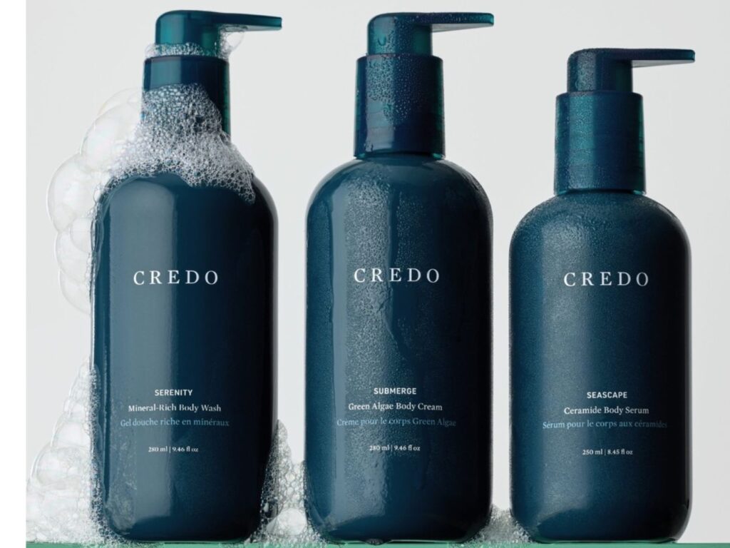 Three marine blue colored bottles of body care products on a shelf, with soap suds around them, with the word CREDO on each