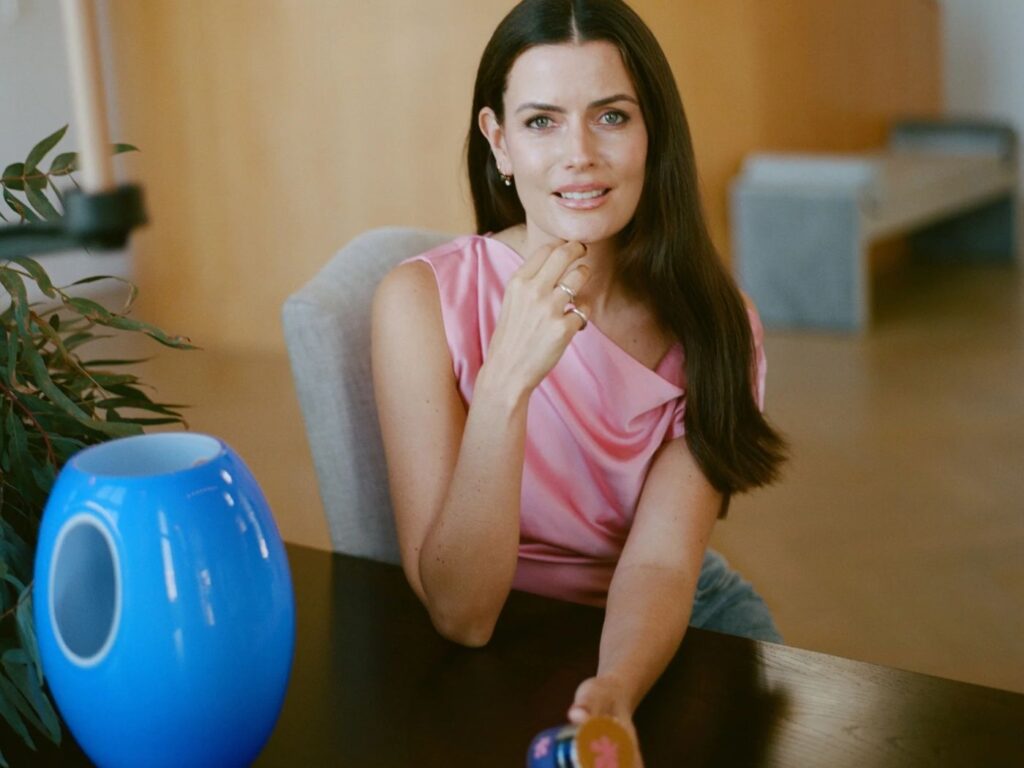 Woman seated at a desk in an office, wearing a pink sleeveless blouse, with a blue vase next to her, and a plant, one arm outstretched and holding a blue jar of face cream.
