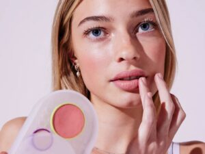 Model holds a makeup compact with peachy cream colour, which she is applying to her lips with her fingertips