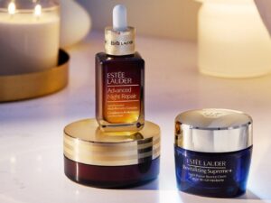 Three Estée Lauder skin care products, including a bottle of Night Repair serum, on a table next to a candle and a glowing lamp.