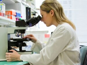 Woman in a white lab coat in a lab office, looking down a microscope